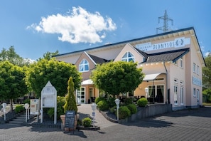 A hotel with a sign reading 'DIONYSOS' and a well-maintained outdoor area with trees and a paved walkway. - HOTEL DIONYSOS (Ransbach-Baumbach)