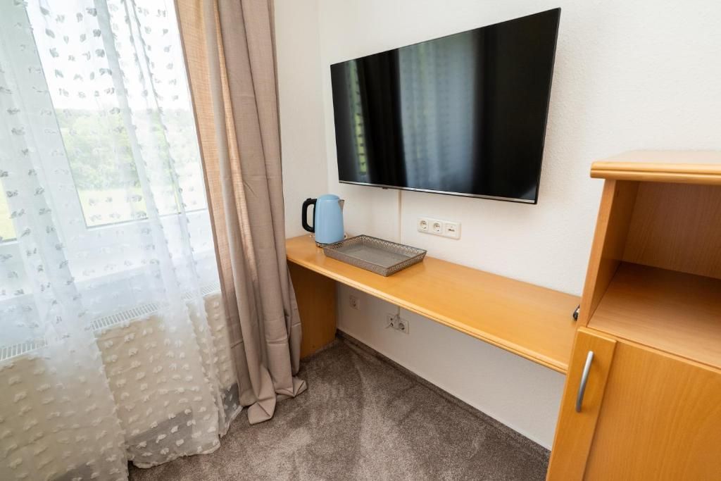 Una habitación con una televisión de pantalla plana montada en la pared, un escritorio de madera con un hervidor y una ventana con cortinas de gasa.