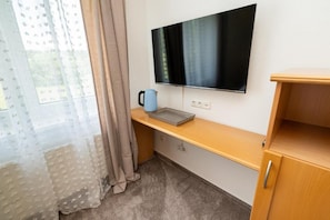 A room with a flat-screen TV mounted on the wall, a wooden desk with a kettle, and a window with sheer curtains. - HOTEL DIONYSOS (Ransbach-Baumbach)