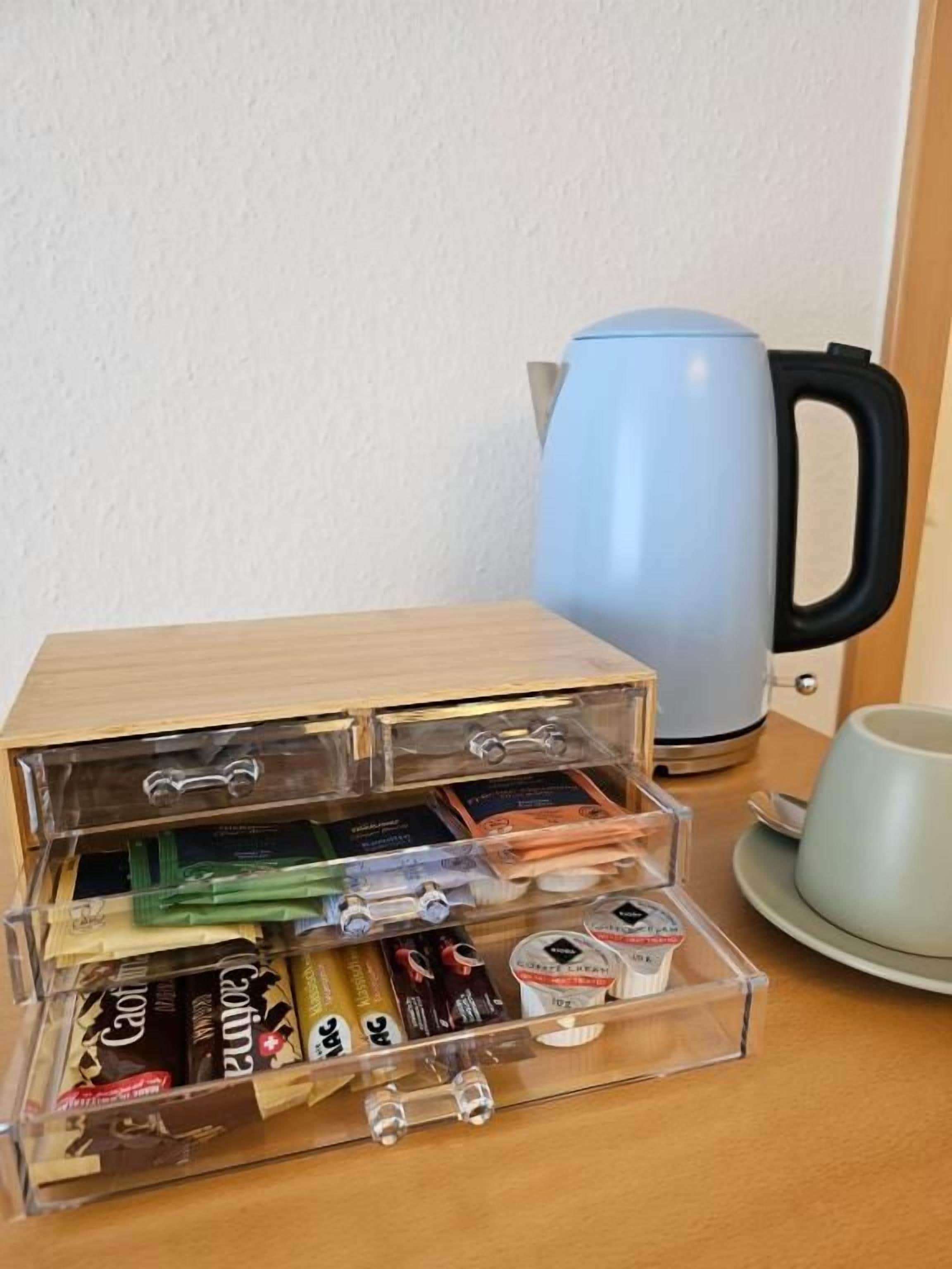 Una mesa de madera con un recipiente transparente que contiene varios objetos, una tetera azul y una taza azul claro sobre un plato.
