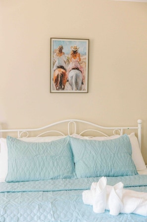 A bed with a quilted blue cover, white pillows, and a framed painting of two horses on the wall. - Mabelle Estates | Historic Mansion • Pool • Farm (Gainesville)