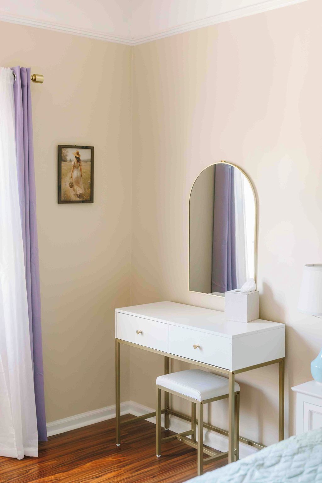 A bedroom with a white vanity, a mirror, a small stool, a painting on the wall, and a curtain.
