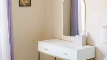 A bedroom with a white vanity, a mirror, a small stool, a painting on the wall, and a curtain.