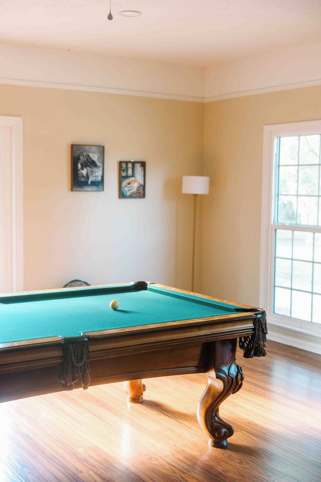 A pool table in a room with wooden flooring, a lamp, and two framed artworks on the wall.