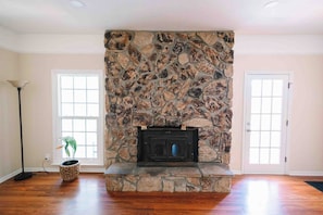 A room with a stone fireplace, hardwood flooring, and two windows. - Mabelle Estates | Historic Mansion • Pool • Farm (Gainesville)
