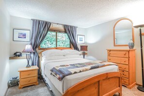 A bedroom with a bed, a wooden dresser, a mirror, a nightstand, and a window with curtains. - Snowater Forest Nook (Deming)