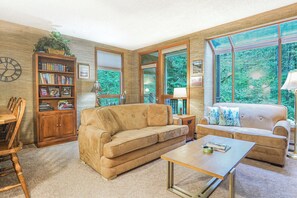 A living room with a beige sofa, a coffee table, a bookshelf, and large windows with a view of greenery. - Snowater Forest Nook (Deming)