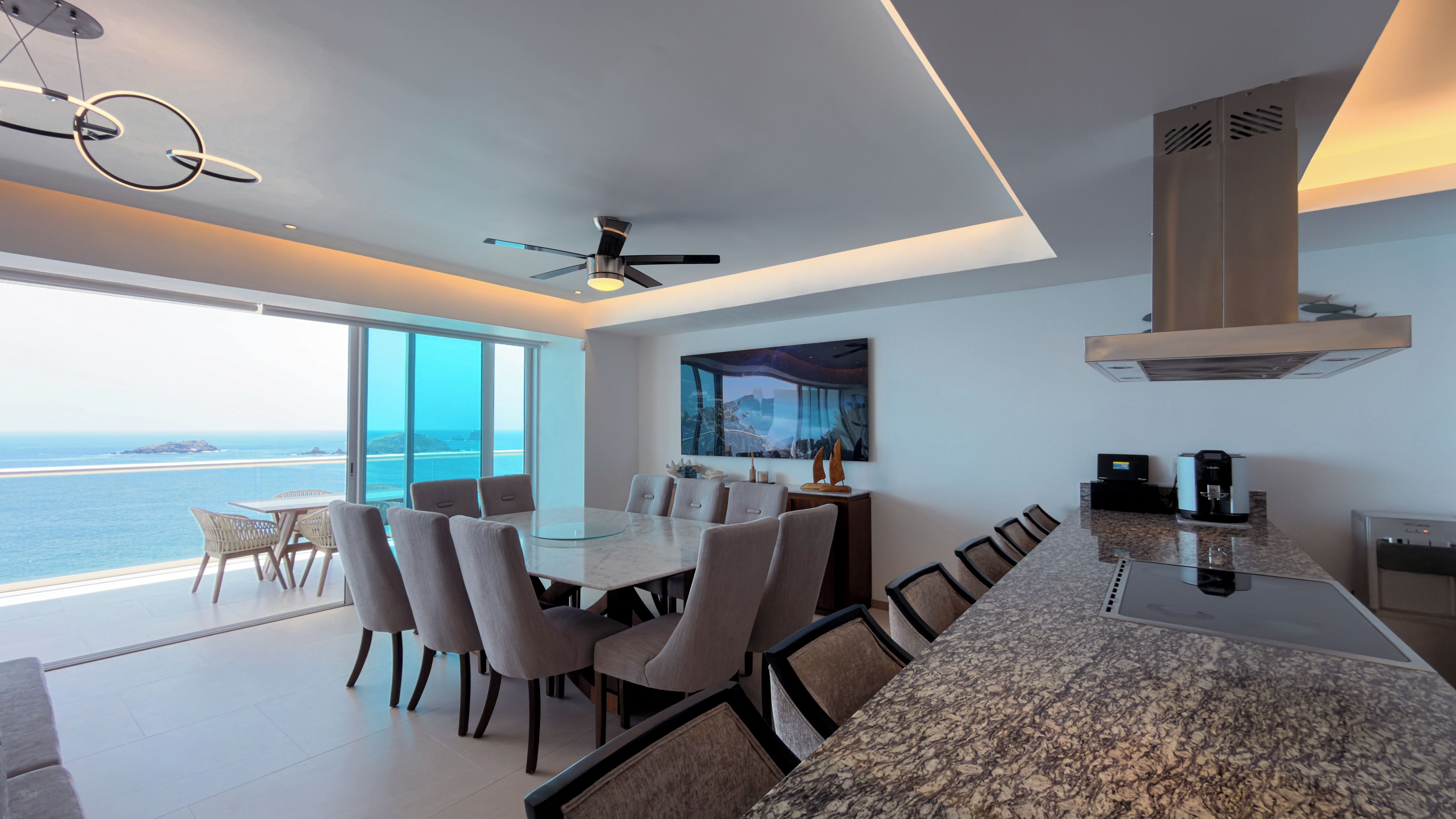 A modern kitchen with a granite countertop, a dining area with a glass table, and a view of the ocean.