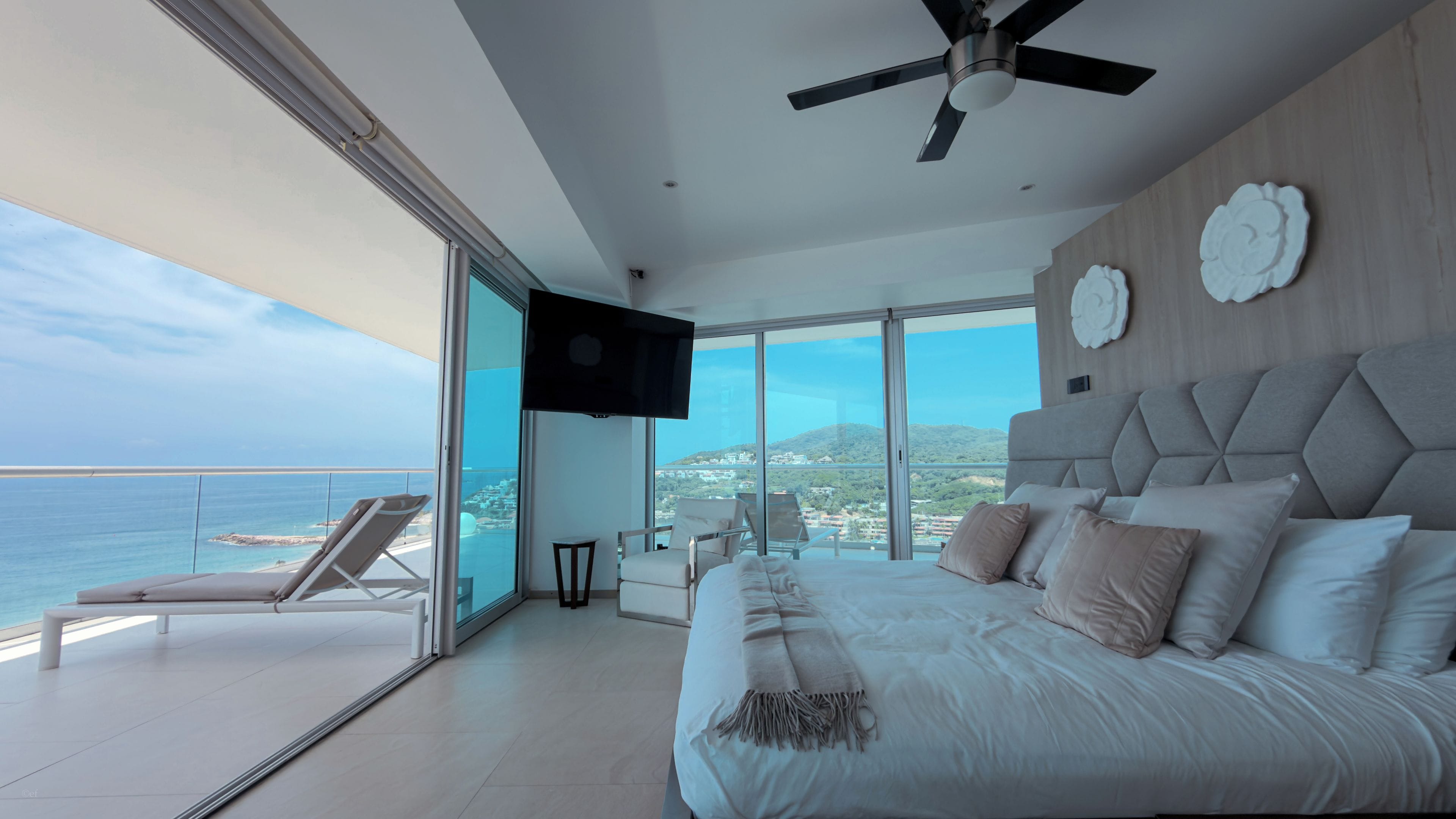 Une chambre avec un grand lit, un ventilateur de plafond et une télévision. Elle dispose d’un balcon avec vue sur la mer.