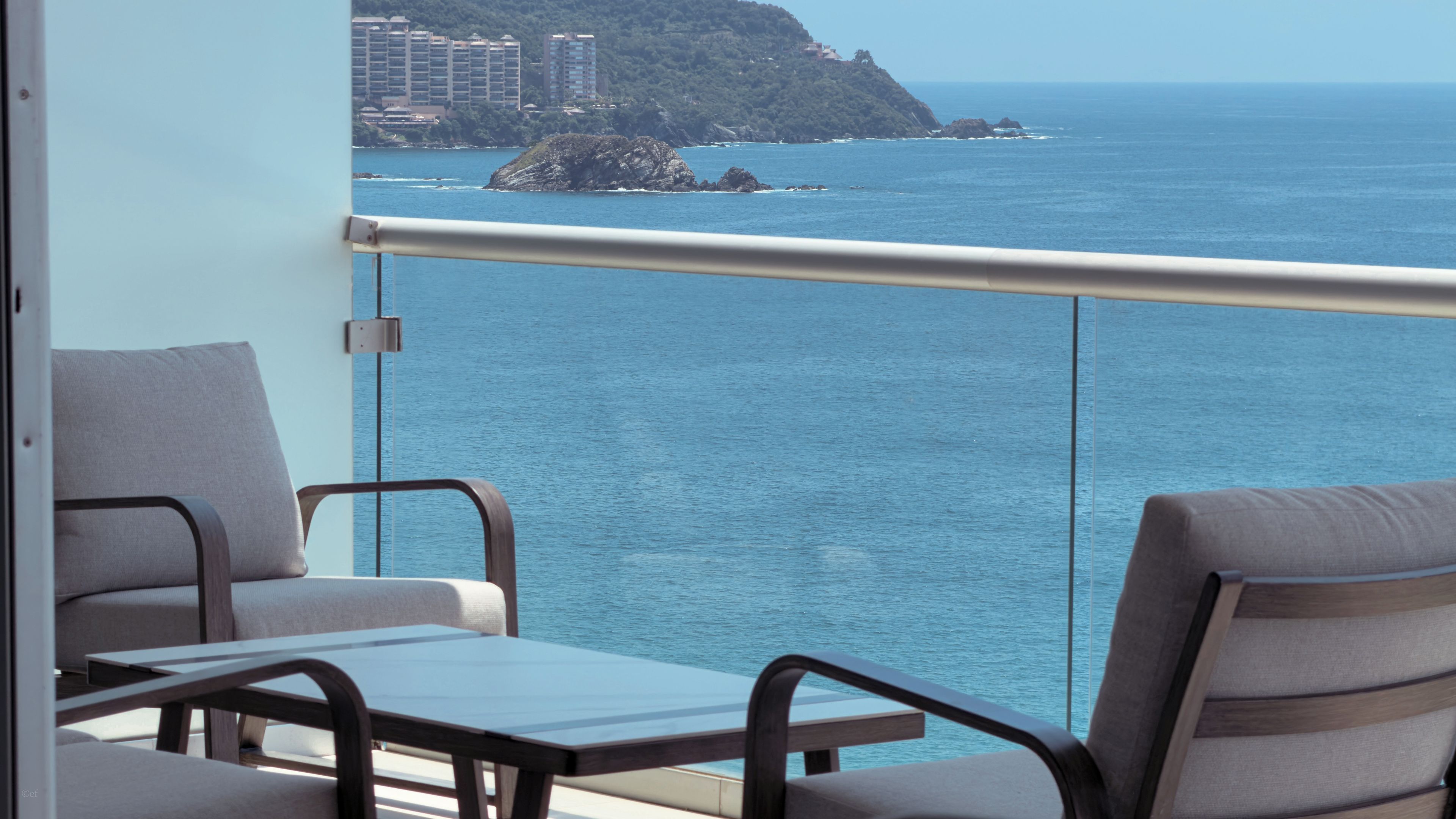 Un balcón con dos sillas y una mesa, con vistas a la costa, edificios y afloramientos rocosos.