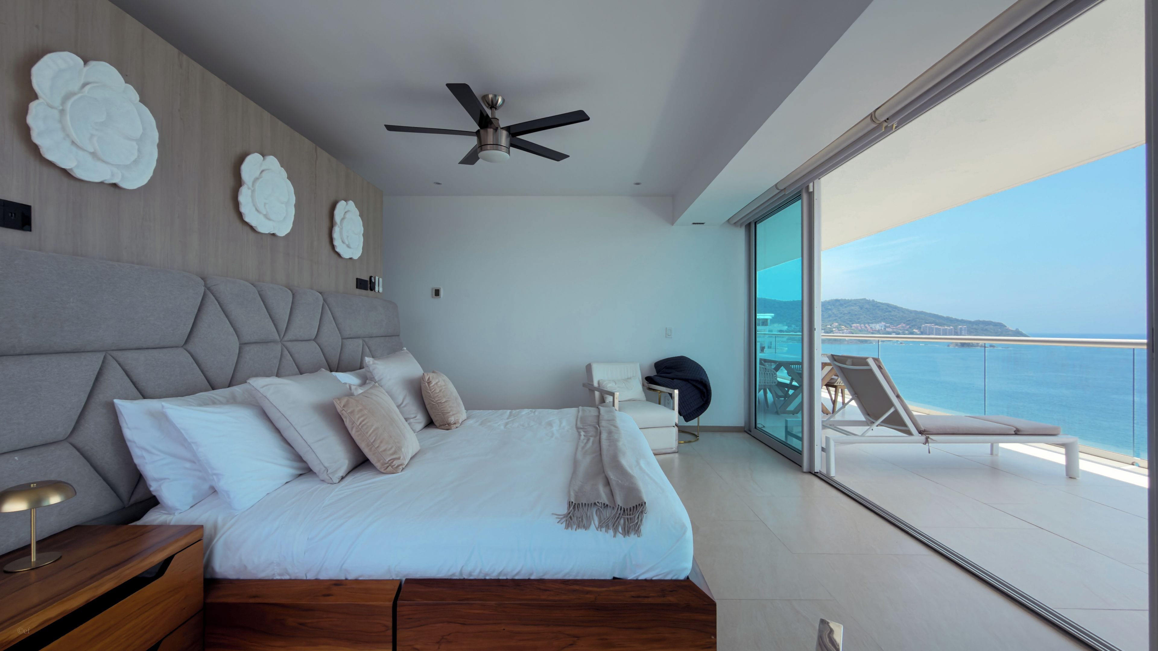 A modern bedroom with a bed, a ceiling fan, a nightstand, a chair, and a view of the sea.