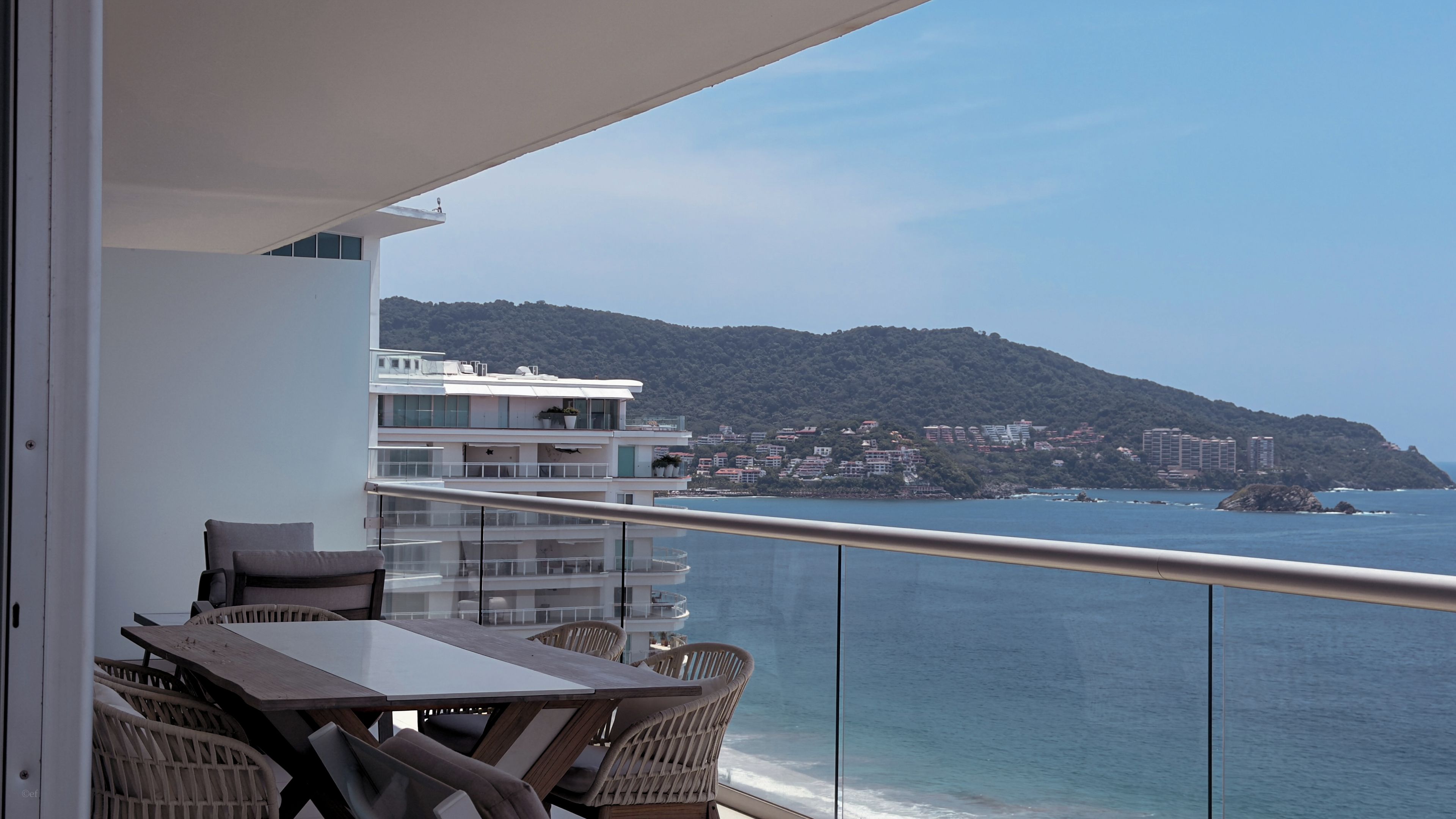 A balcony with a table and chairs, overlooking a coastal area with buildings and a hill.
