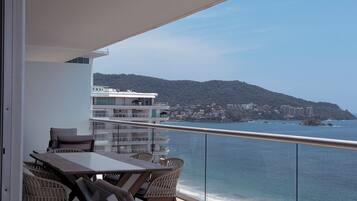 A balcony with a table and chairs, overlooking a coastal area with buildings and a hill.