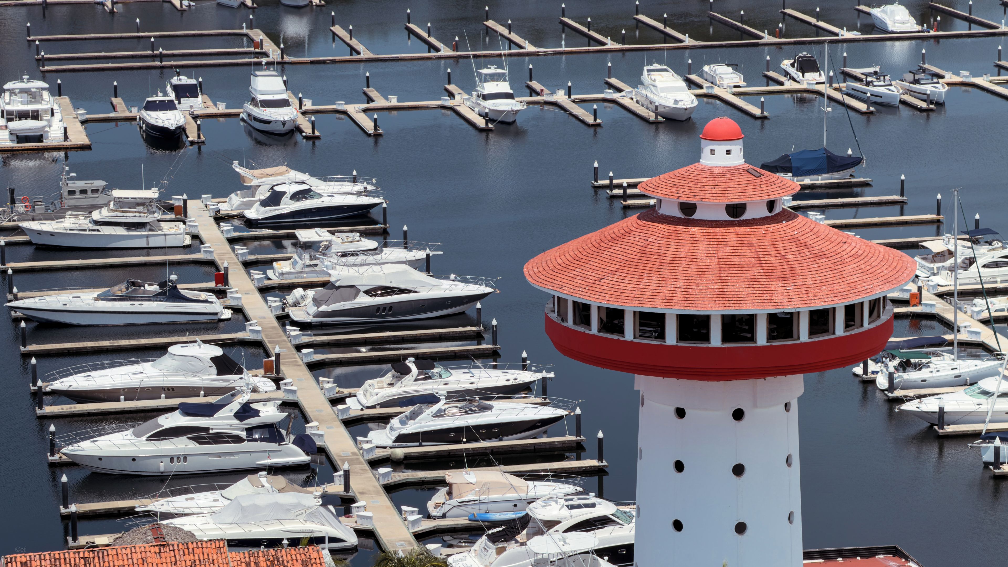 A marina with multiple boats docked at various piers, a prominent lighthouse structure, and a clear view of the water.