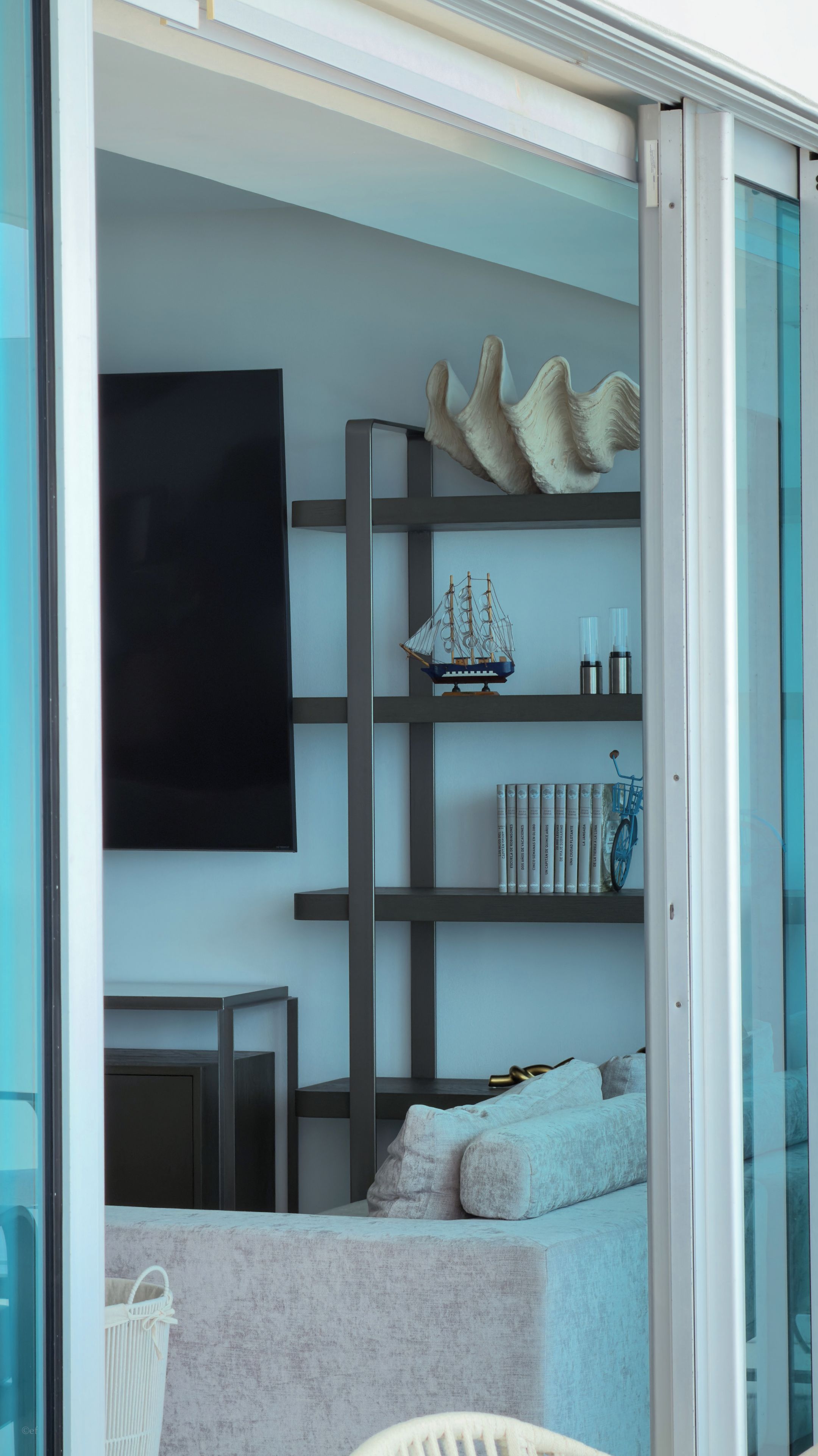 A modern interior with a flat-screen TV mounted on the wall, a bookshelf with decorative items, and a seating area with a sofa and cushions.