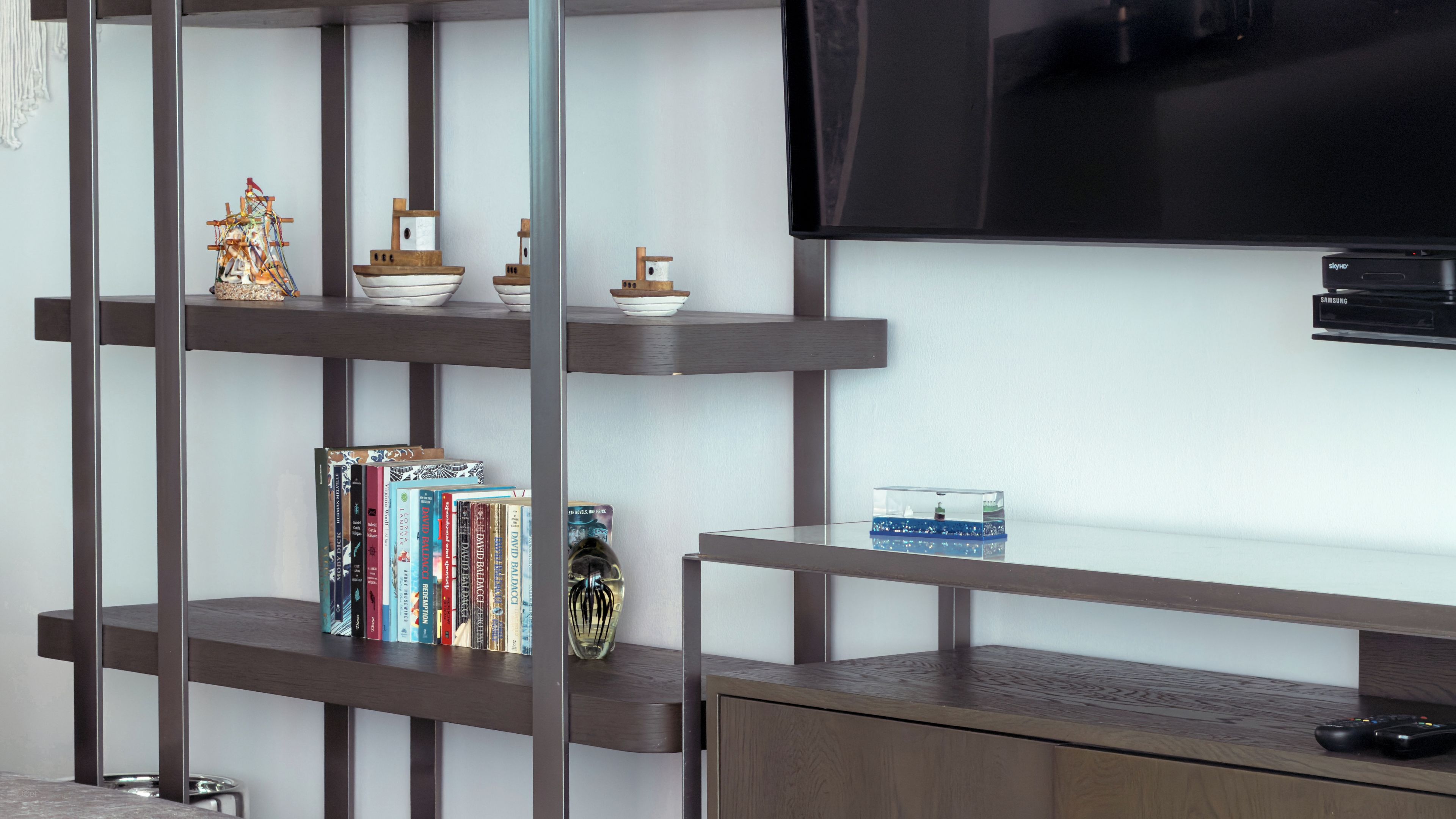 Una habitación con una televisión montada en la pared, un armario de madera y una estantería con libros y objetos decorativos.