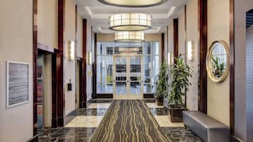 A modern hotel lobby with a marble floor, a bench, potted plants, and a large glass door leading to the reception area.