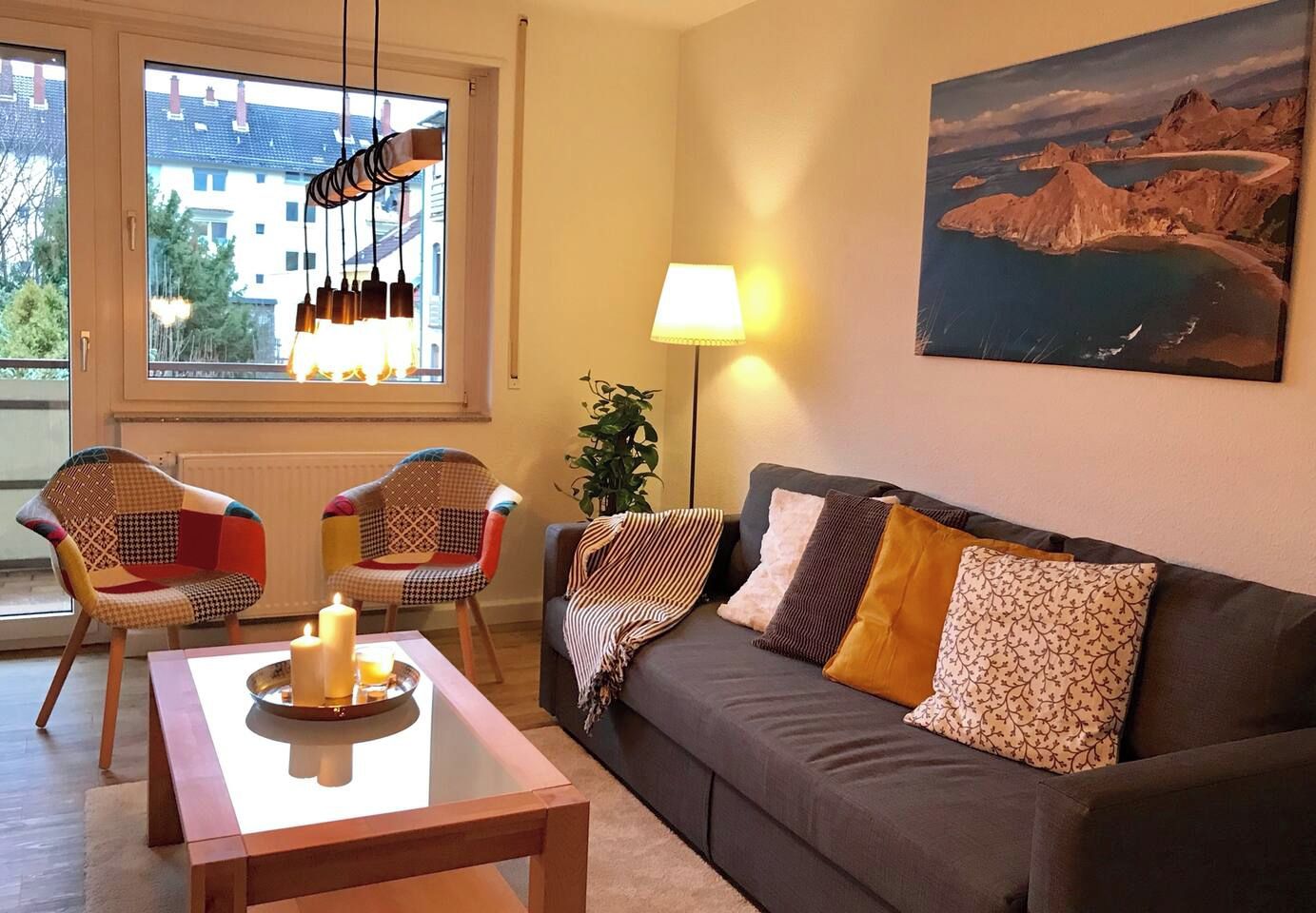 A living room with a sofa, two chairs, a table with candles, a lamp, and a painting of a coastal landscape.