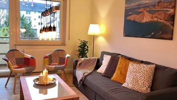 A living room with a sofa, two chairs, a table with candles, a lamp, and a painting of a coastal landscape.