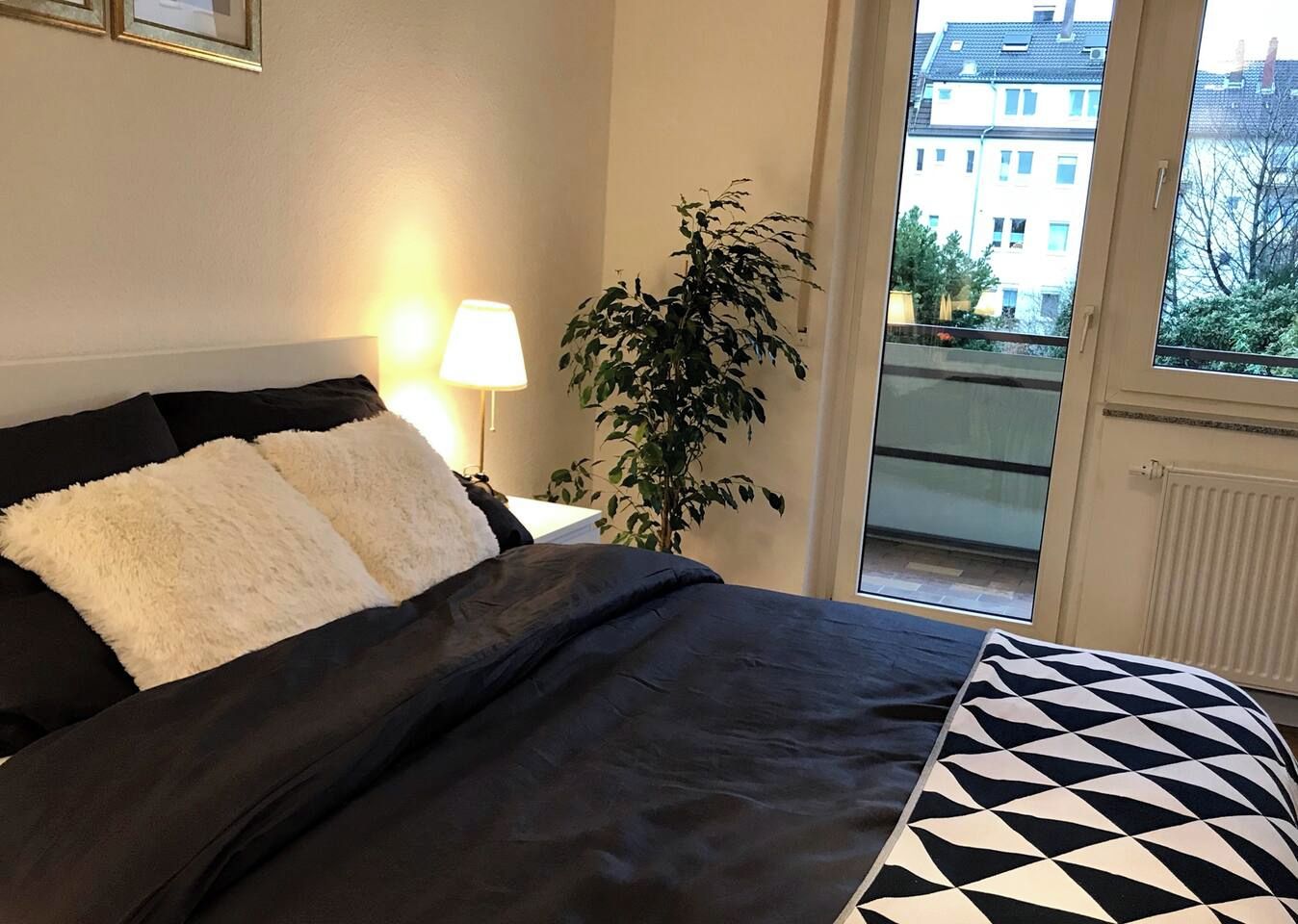 Une chambre à coucher avec un lit, une lampe, une plante en pot et une vue sur des immeubles par la fenêtre.