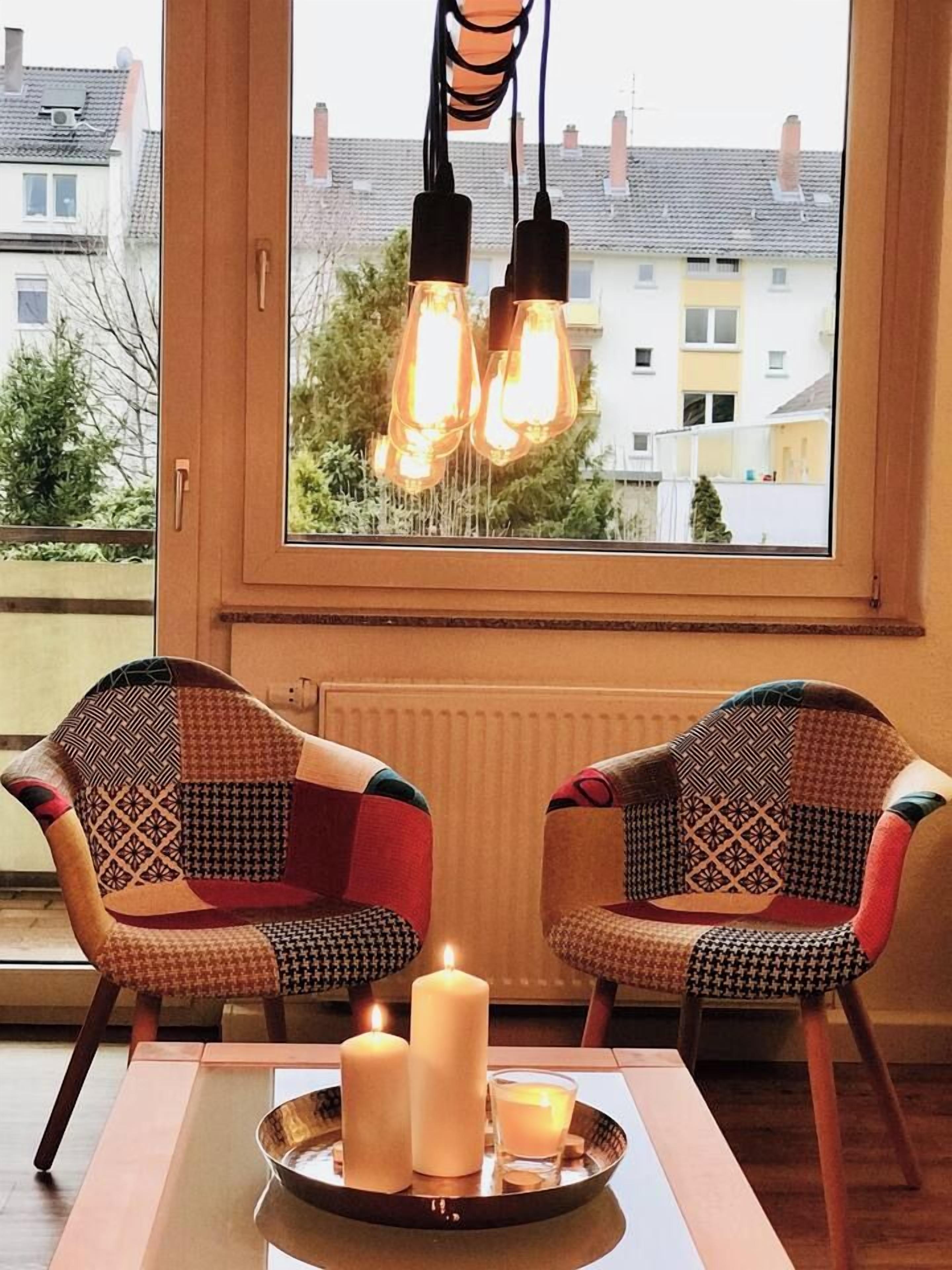 Ein Zimmer mit zwei gemusterten Sesseln, einem Tisch mit Kerzen und Blick durch ein Fenster mit hängenden Lampen.