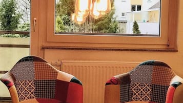 Ein Zimmer mit zwei gemusterten Sesseln, einem Tisch mit Kerzen und Blick durch ein Fenster mit hängenden Lampen.