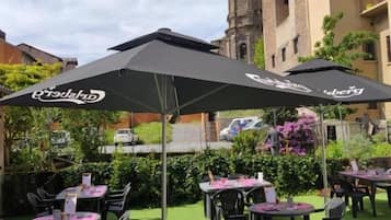 Un'area pranzo all'aperto con tavoli e sedie, ombrelloni e un edificio con una torre dell'orologio sullo sfondo.