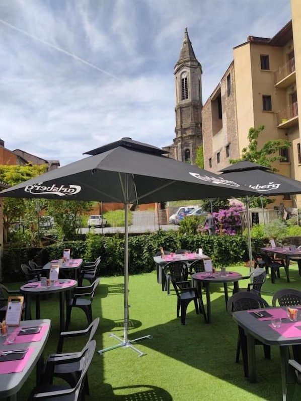 An outdoor dining area with tables and chairs, umbrellas, and a building with a clock tower in the background.