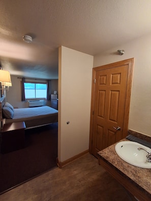A hotel room with a bed, a bathroom with a sink, and a window. - Cyclone Lodge By Fresh Stay (Harlan)