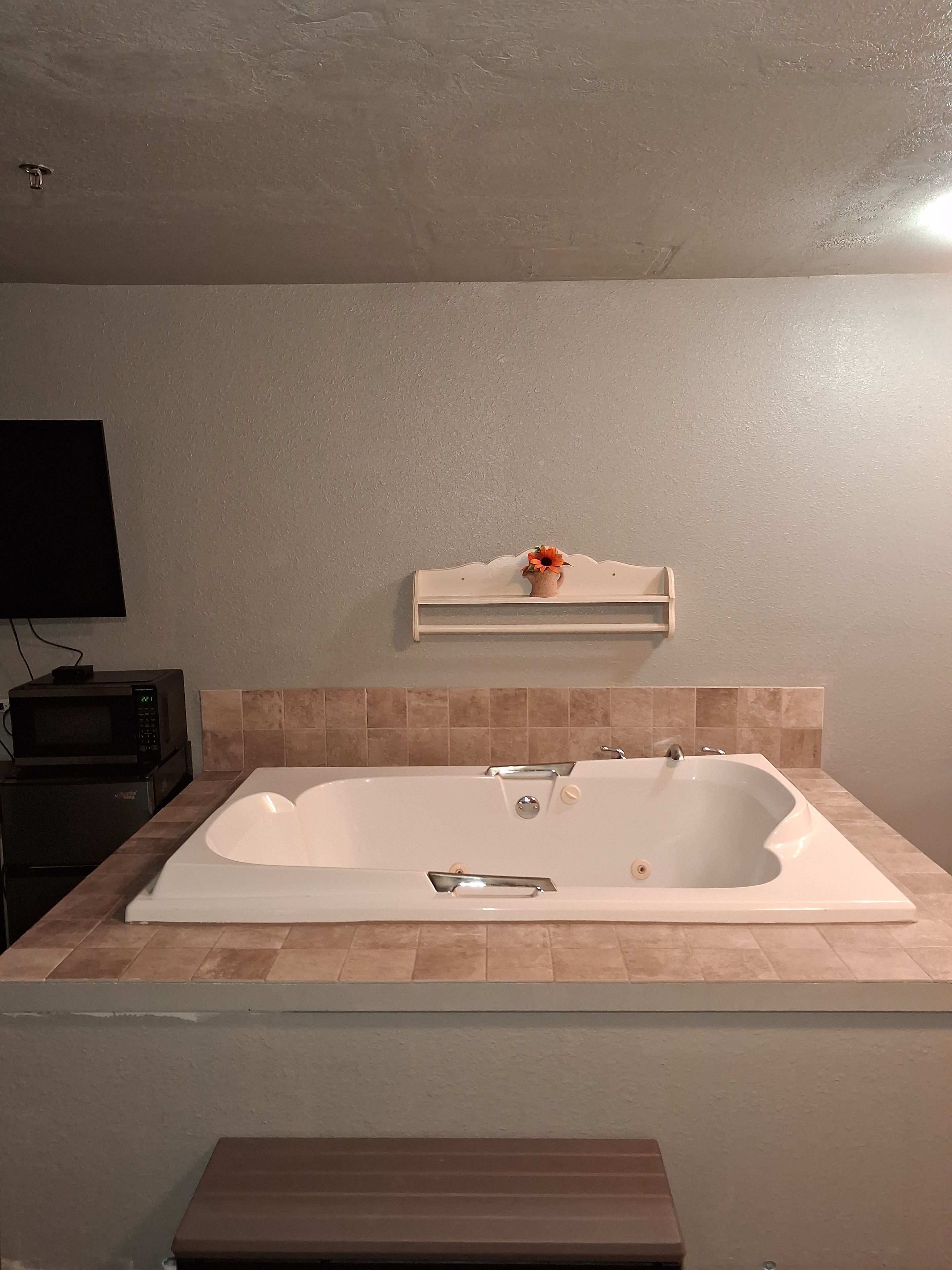 A bathroom with a white bathtub, a small shelf with a flower, and a microwave on the countertop.