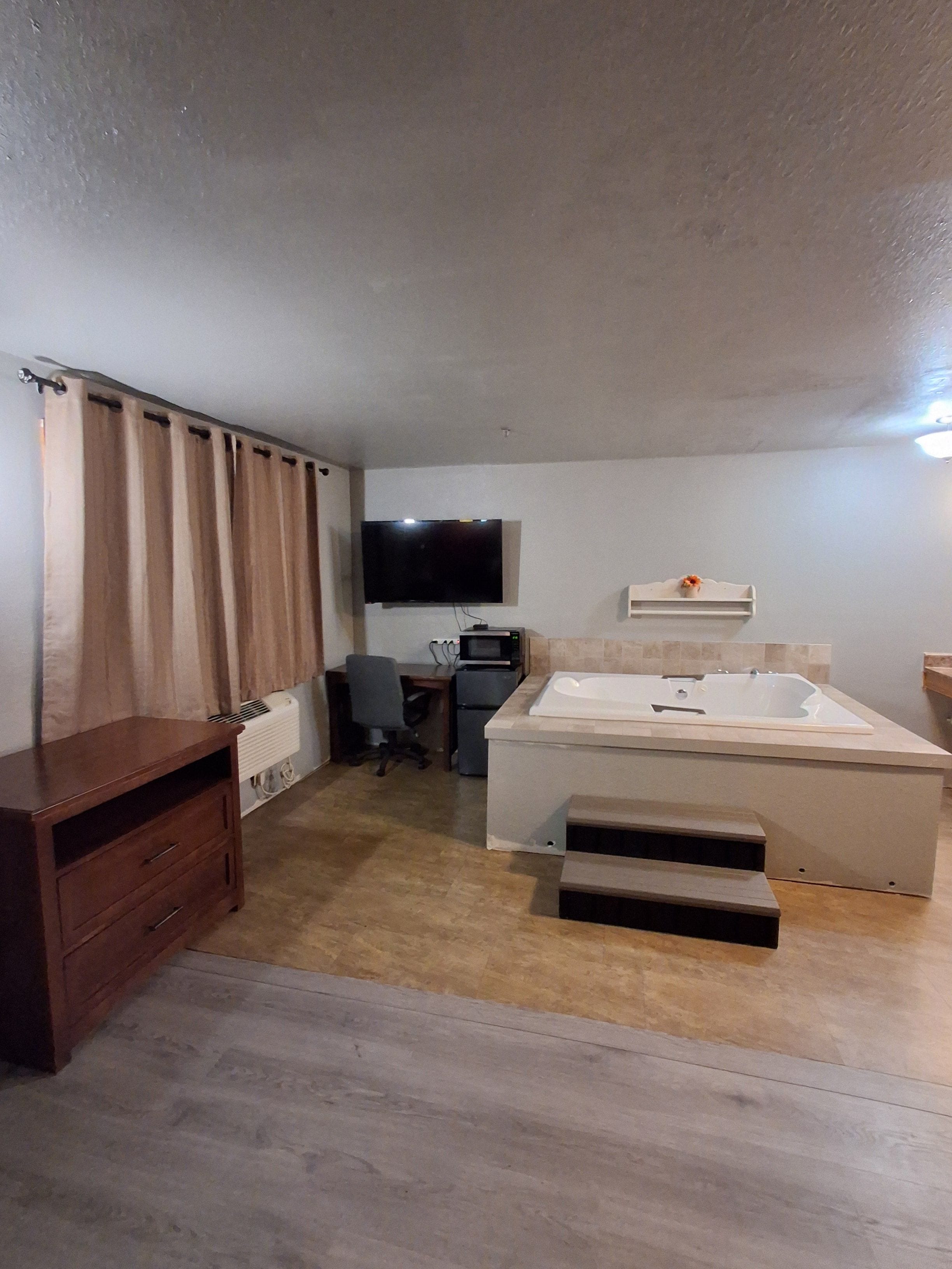 A compact room with a flat-screen TV, a small desk with a chair, a built-in bathtub, and a wooden cabinet.