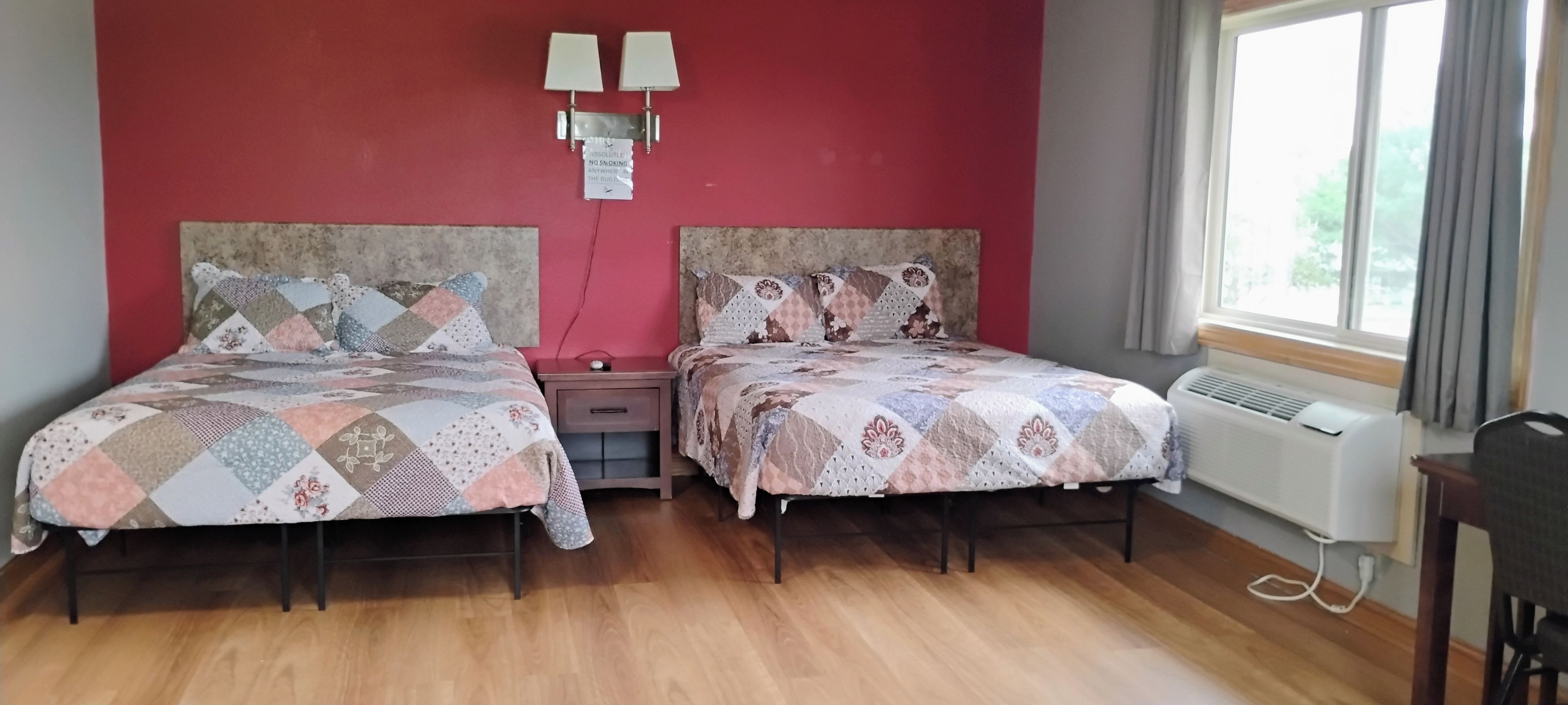 A room with two beds, a nightstand, a wall-mounted light fixture, and a window with curtains.