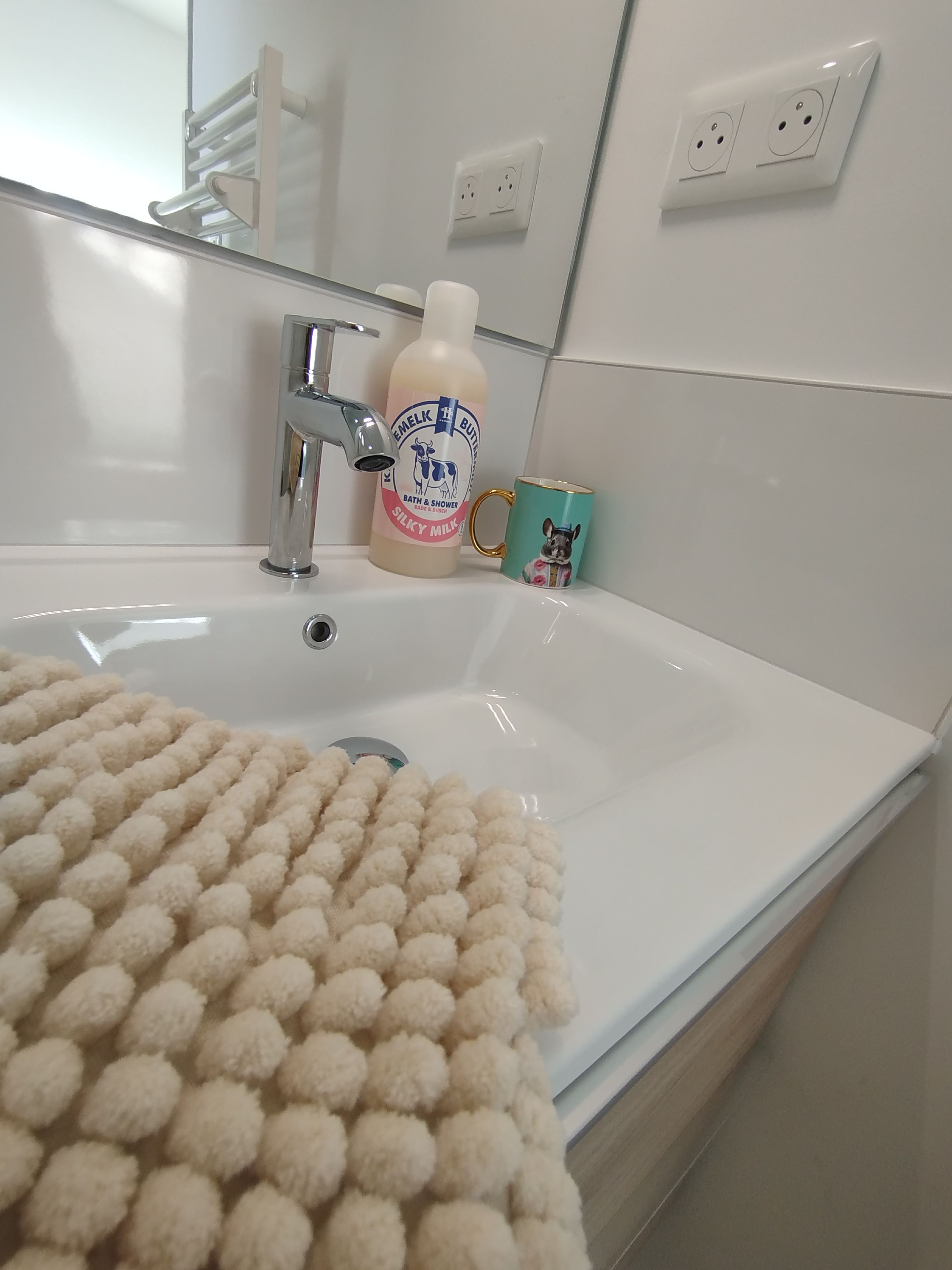 A bathroom with a white sink, a bottle of bath & shower soap, a mug, and a fluffy beige bath mat.