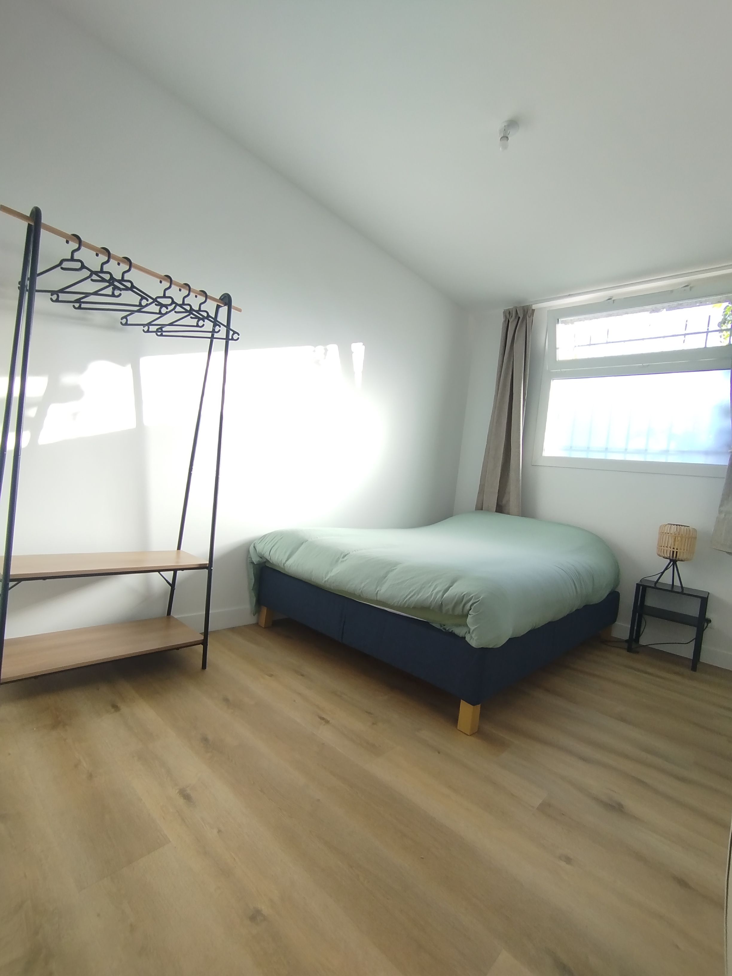 A bedroom with a bed, a wooden floor, a window with curtains, and a wall-mounted shelf.