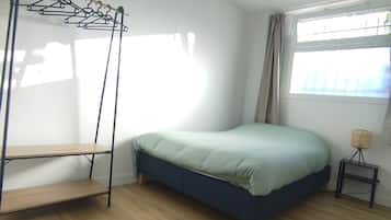 A bedroom with a bed, a wooden floor, a window with curtains, and a wall-mounted shelf.