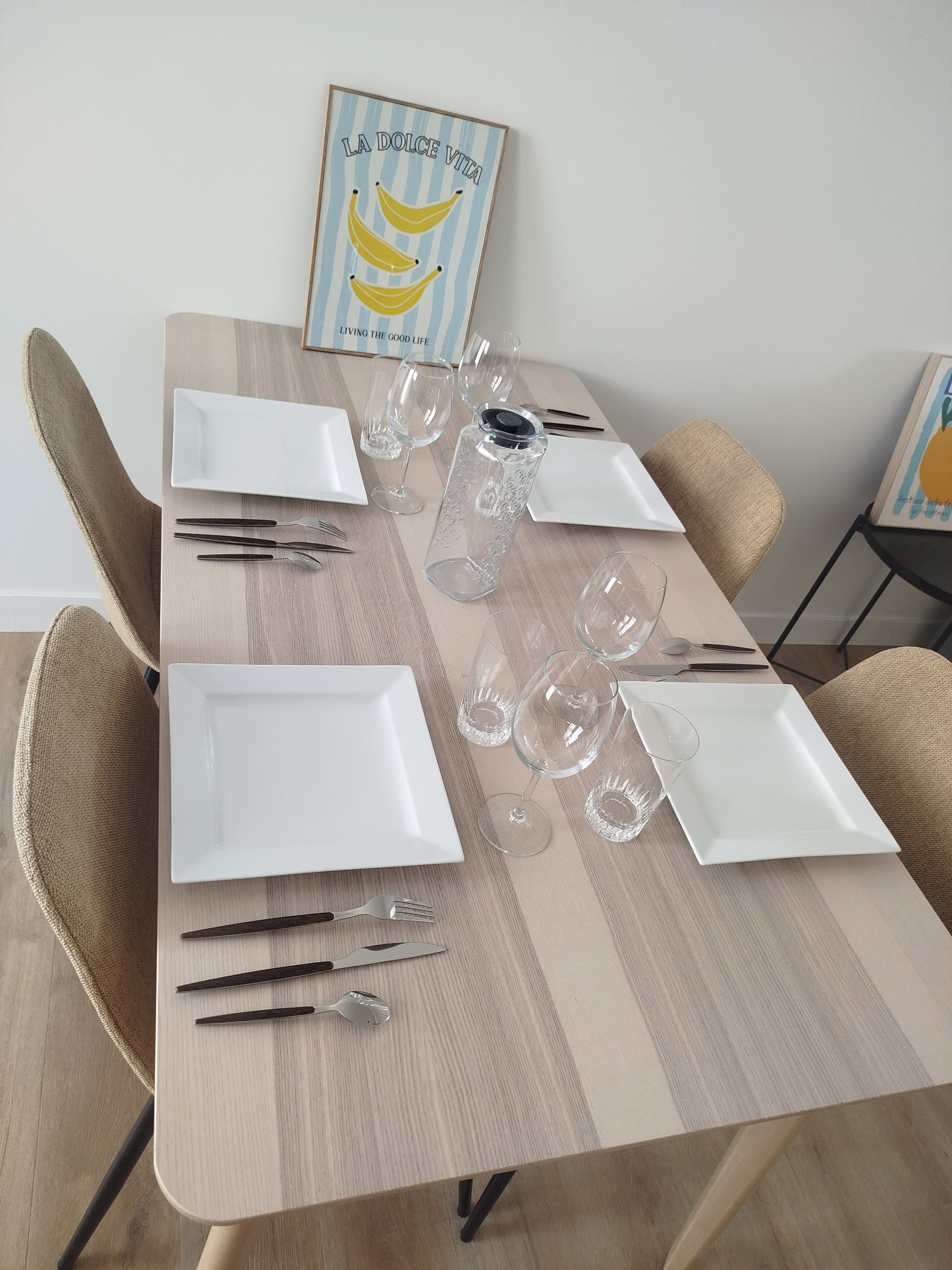 Een eettafel gedekt met witte borden, bestek en wijnglazen, met daarboven een lijst met een foto.