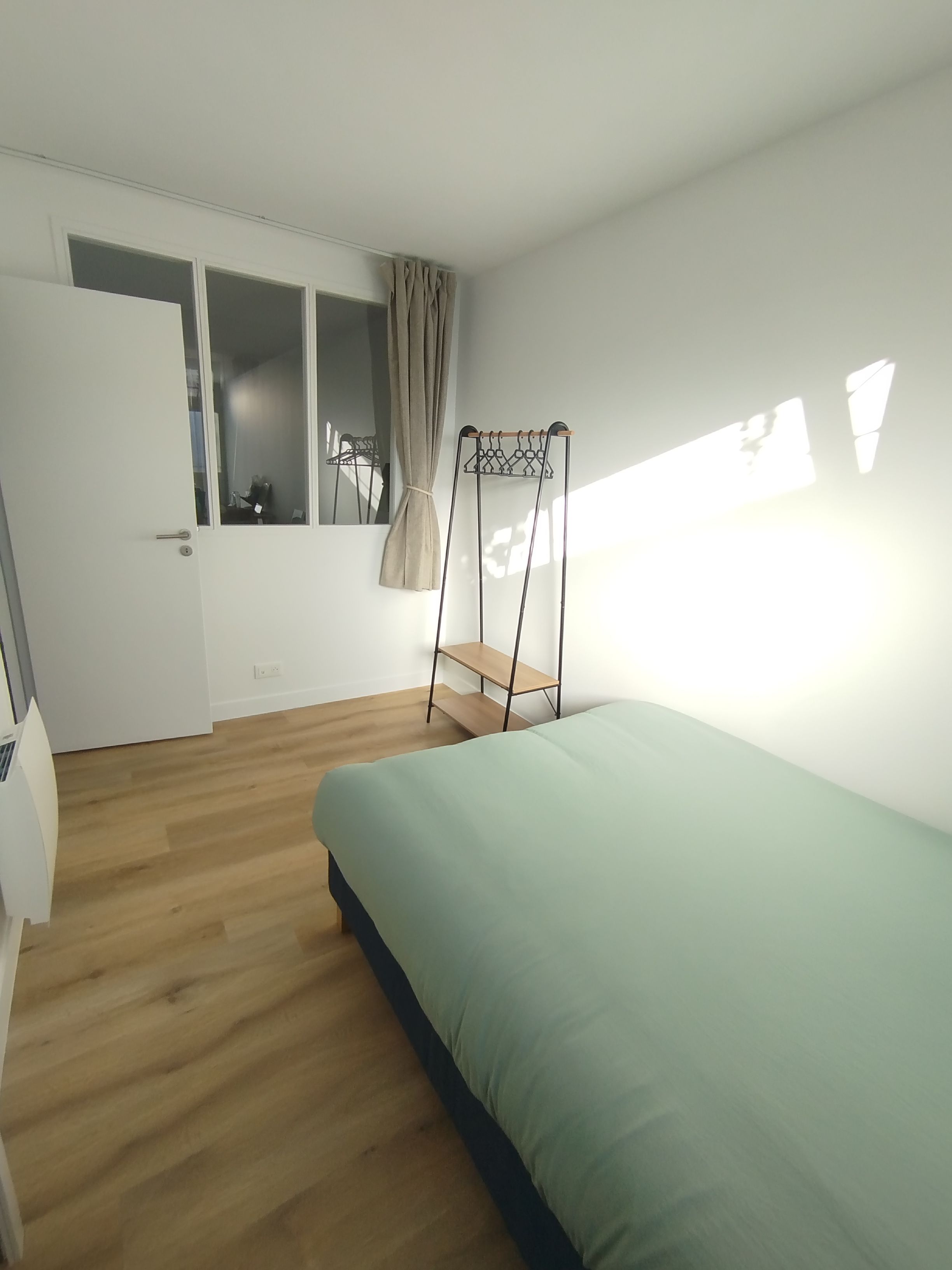 A room with a bed, a window with curtains, a door, and a small shelf.