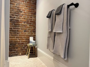 Ein Badezimmer mit Backsteinwand, einem hölzernen Hocker mit Toilettenartikeln und zwei Handtüchern, die an einem Wandhaken hängen.