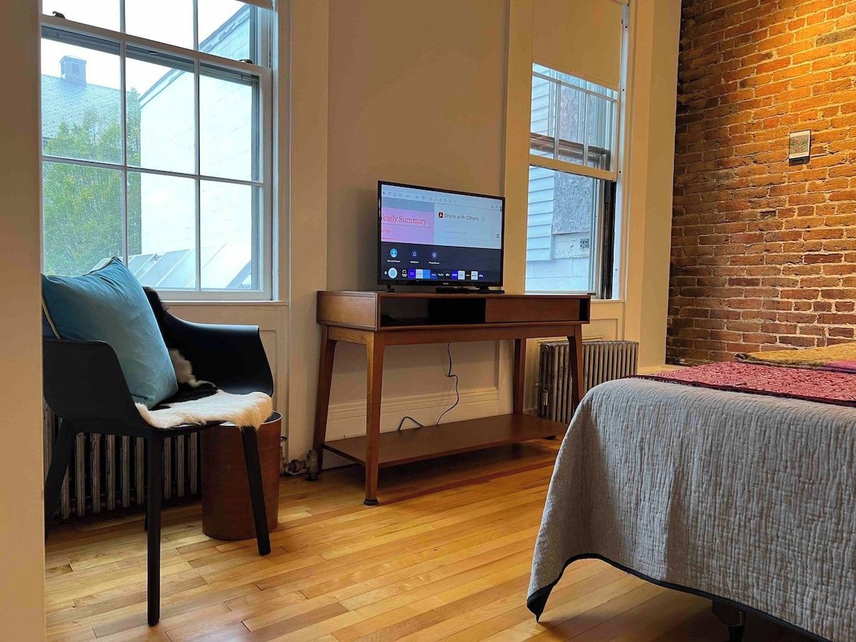 A room with a wooden floor, a bed with a grey bedspread, a black chair with a blue cushion, a wooden desk with a computer, and a brick wall.