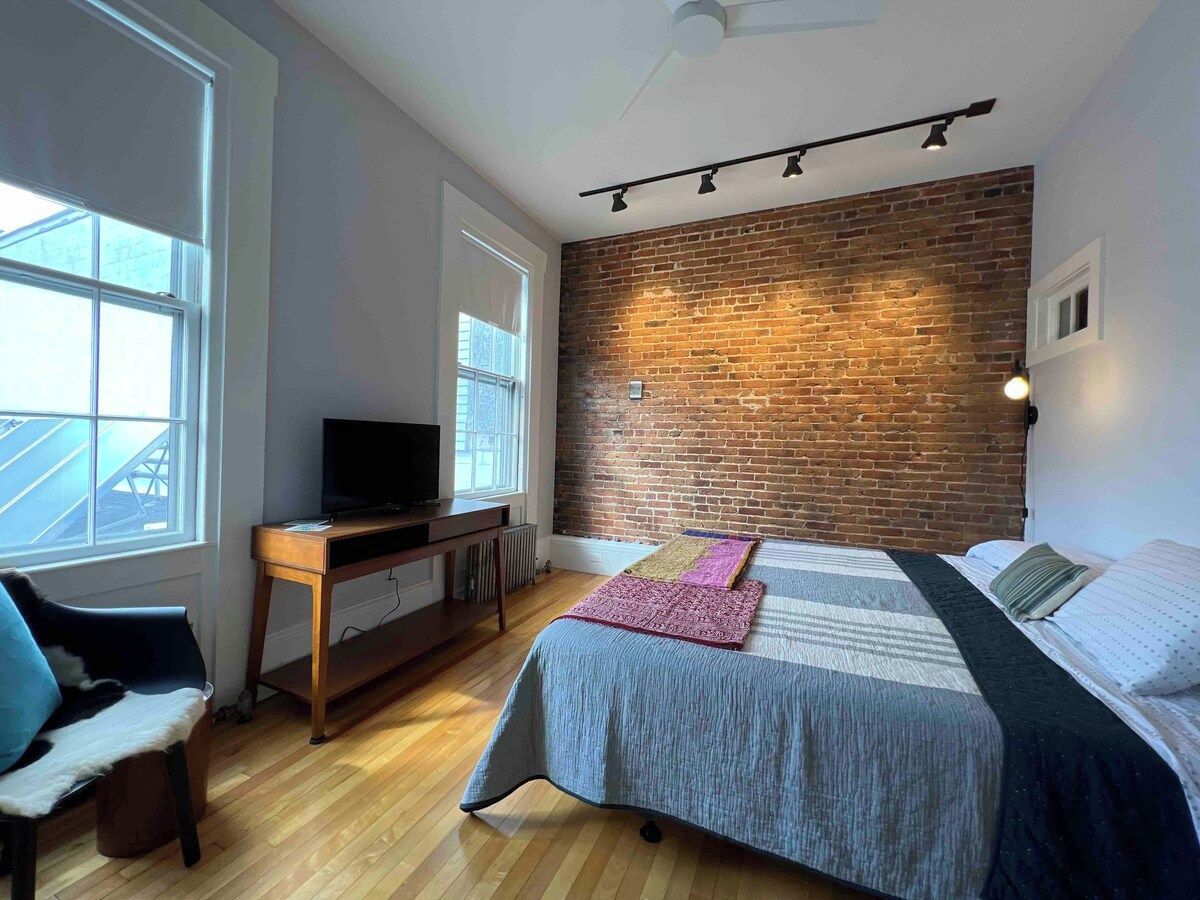 Quarto com parede de tijolos, cama com várias almofadas, escrivaninha com televisão e janela com vista para um edifício.