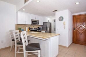 A kitchen with white cabinets, a stainless steel refrigerator, a microwave, and a sink. There are two white chairs with black cushions. - Cozy Ground Level Condo w/ Pool | Princesa E-103 (Puerto Peñasco)