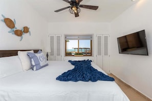 A hotel room with a bed, a ceiling fan, a TV, and a window with a view of a beach and a pool. - Cozy Ground Level Condo w/ Pool | Princesa E-103 (Puerto Peñasco)