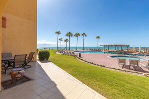 A pool area with lounge chairs, a small table, and a view of the ocean. - Cozy Ground Level Condo w/ Pool | Princesa E-103 (Puerto Peñasco)