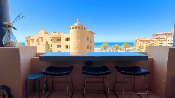 Ein Balkon mit Tisch und drei Stühlen, der Blick auf einen Strand und ein Gebäude mit einer Kuppel bietet.