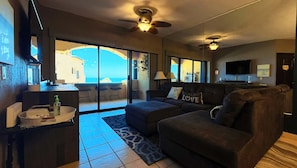 Ein modernes Wohnzimmer mit einer Couch, einem Fernseher und Blick auf das Meer durch große Glasüren.
