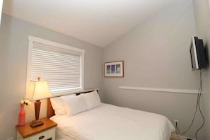 A bedroom with a bed, a nightstand, a lamp, a window with blinds, a wall-mounted TV, and a small framed picture.