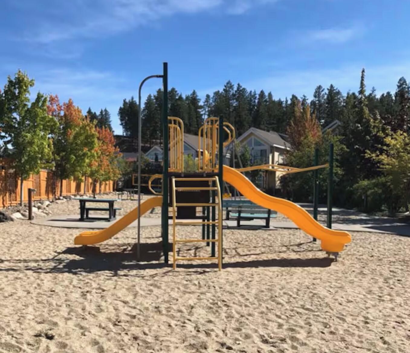 Une aire de jeux avec un toboggan et des structures d’escalade sur un sol sablonneux, entourée d’arbres et de maisons.