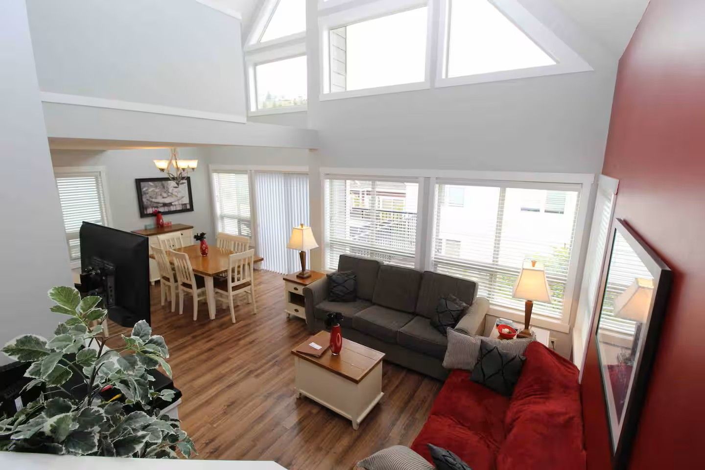 A spacious living area with a dining table, a sofa, and a red couch. There are large windows allowing natural light.