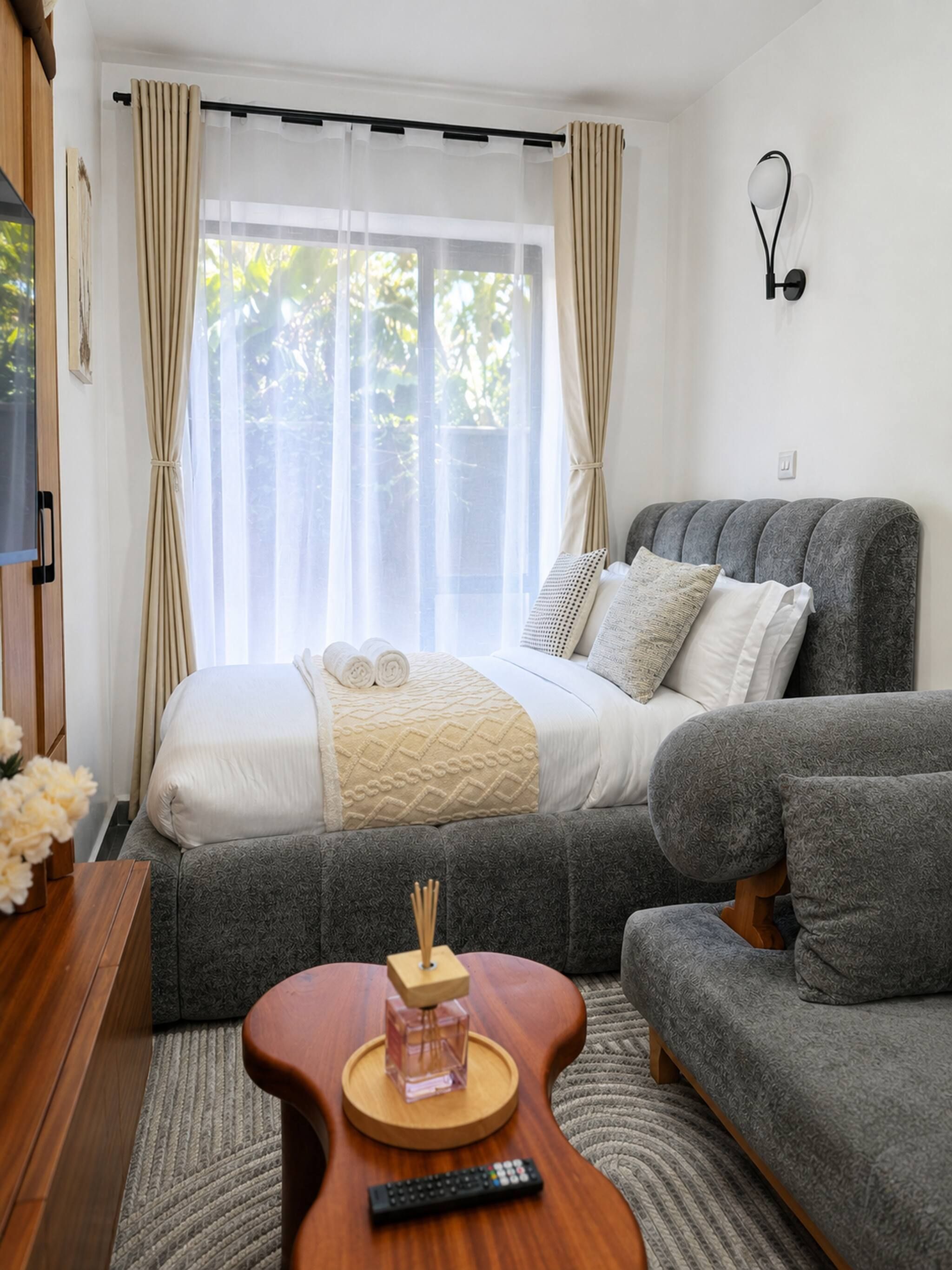 Quarto moderno com cama, sofá revestido de cinza e mesa de cabeceira de madeira com controle remoto.