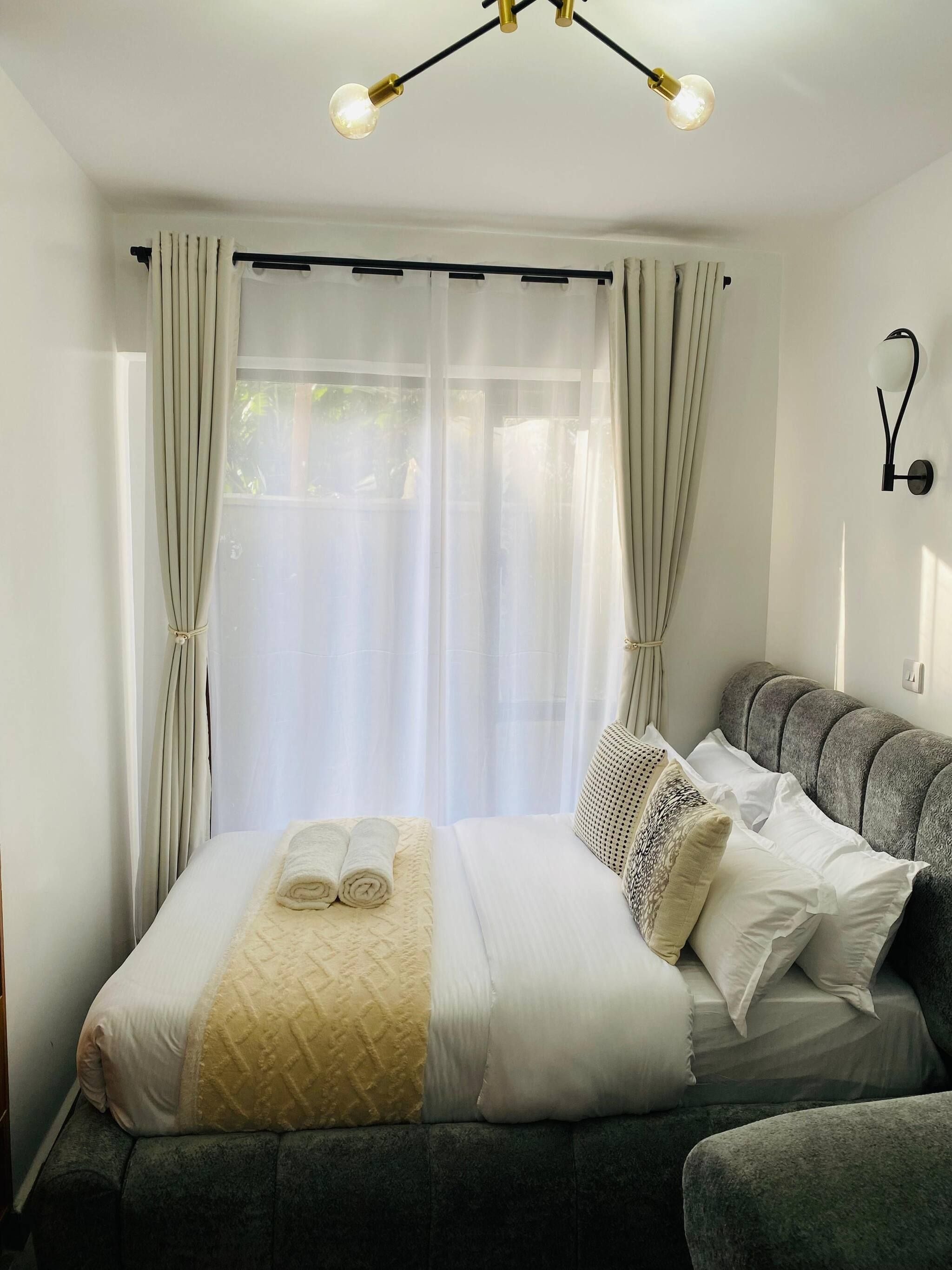 A neatly made bed with a duvet, pillows, and a rolled towel. There is a grey sofa and a wall-mounted light fixture.
